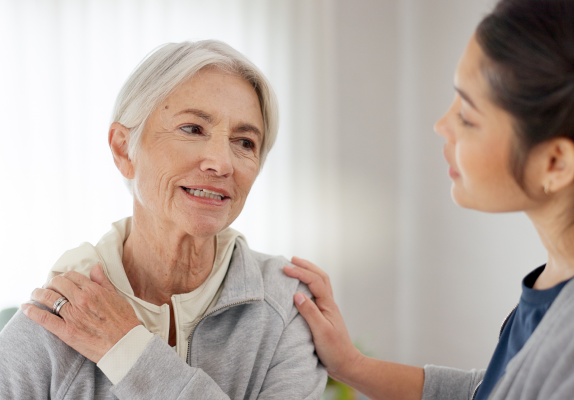 Older woman holding her shoulder talking to younger female nurse.